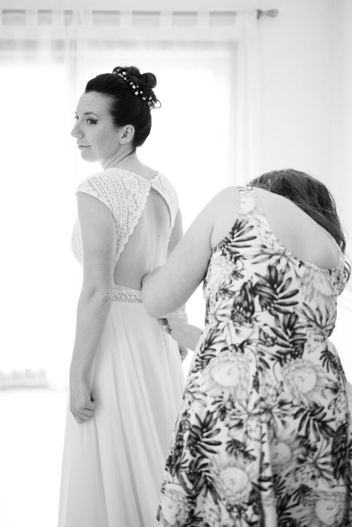 En noir et blanc, une femme ajuste le dos de la robe de la mariée pendant les préparatifs.