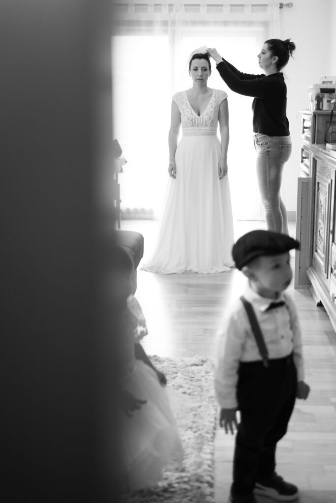 La mariée se fait coiffer près d’une fenêtre, tandis qu’un petit garçon en casquette et bretelles se tient au premier plan.